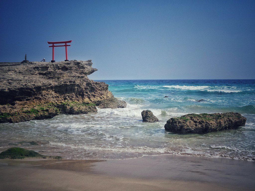 A red torii gate on a rocky outcrop above turquoise sea and a sandy beach